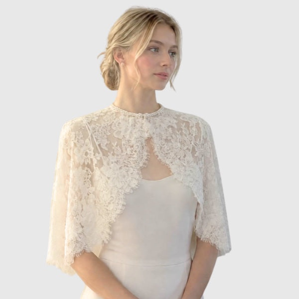 A woman wearing an ivory lace short cape over a matching ivory silk dress with pearl bead neckline.
