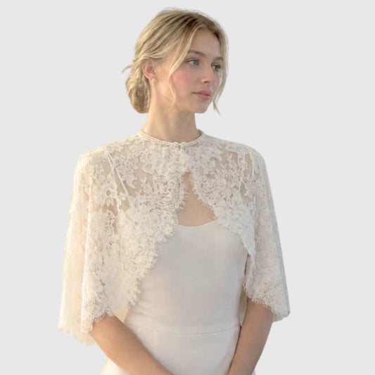 A woman wearing an ivory lace short cape over a matching ivory silk dress with pearl bead neckline.
