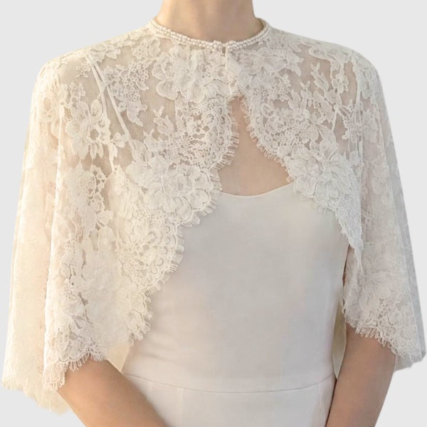 An ivory lace bridal cape for with floral lace patterns and a pearl-trimmed neckline, worn over a matching cream silk dress.